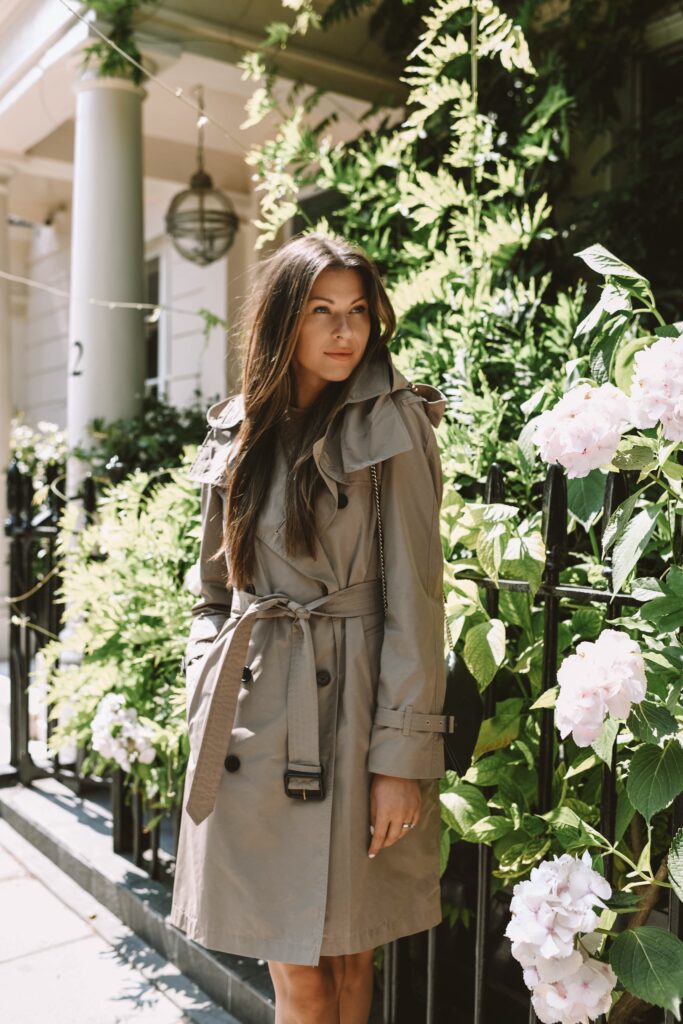 Sieviete stāv pie puķēm ar brūnu ''Burberry'' tranšejas mēteli