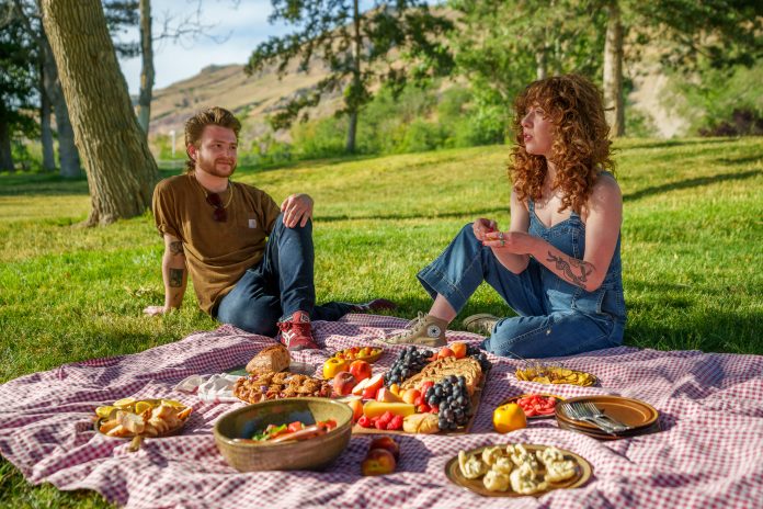 Pikniks dabā, pāris pie uzklāta pleda uz zāles izbauda pikniku.