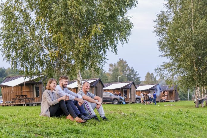 Interesantas ekskursijas Latvijā. Trīs cilvēki sēž zālē pie kempinga mājām.