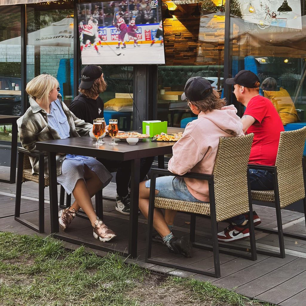 Cilvēki ārā pie galdiņa skatās hokeju un izbauda Maxi Mārupe ēdienu.