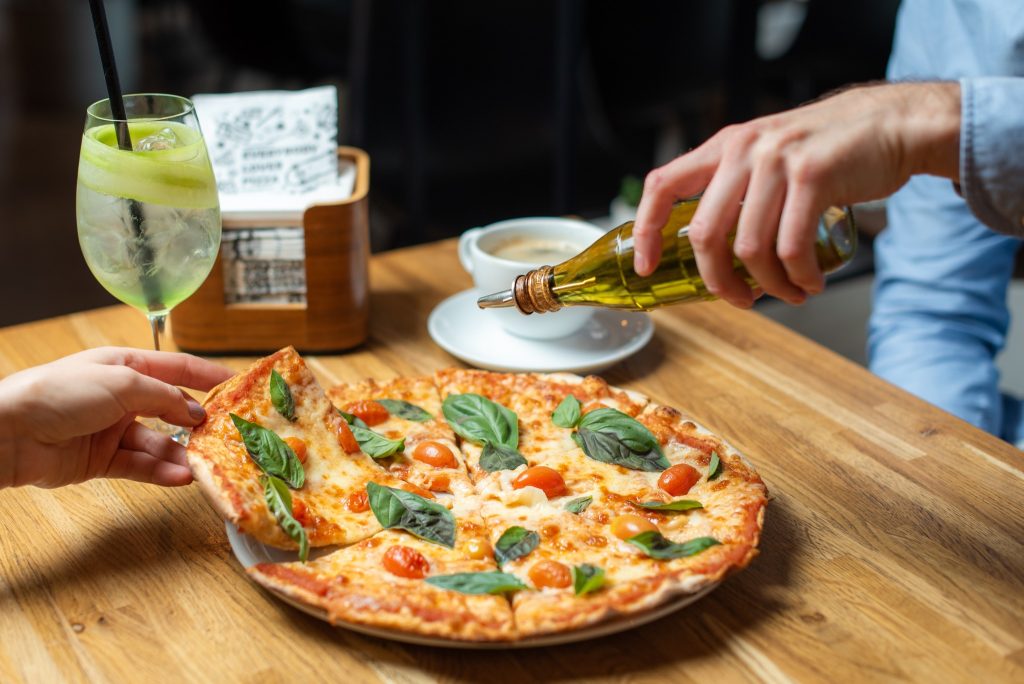 Studio Pizza Cēsīs. Cilvēks lej eļļu uz siera picas ar tomātiem.