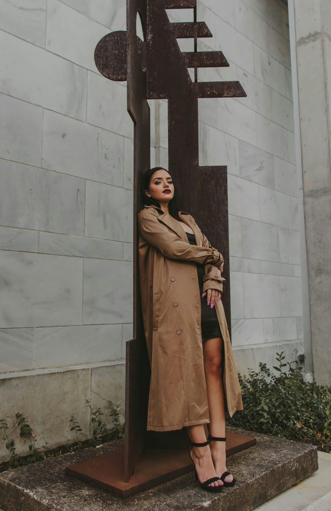 Sieviete brūnā mētelī pozē pie vides objekta