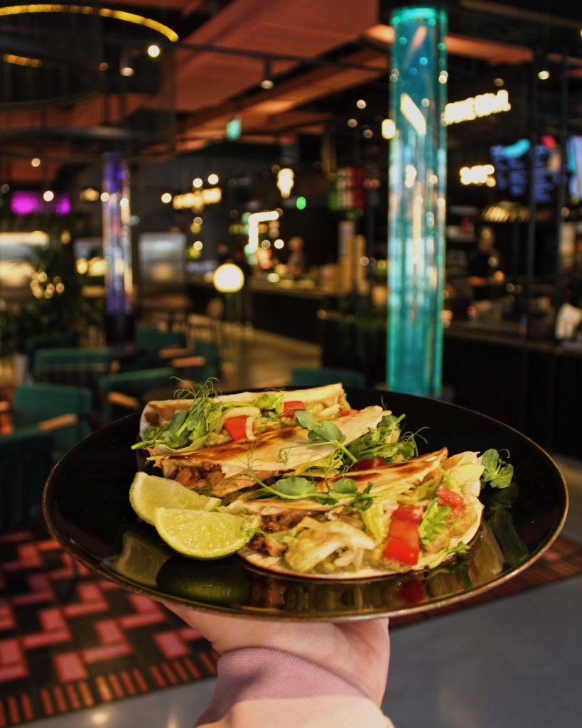 Vīrietis tur šķīvi ar tacos no L-Taco restorāna Burzmā