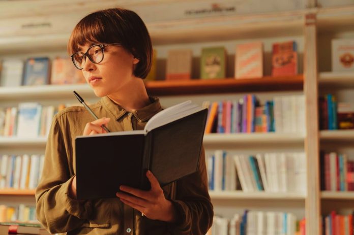 Sieviete ar kladi un pildspalvu bibliotekā